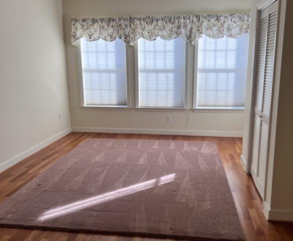 Hardwood living room floor with brown area carpet and 3 windows with natural lighting. 